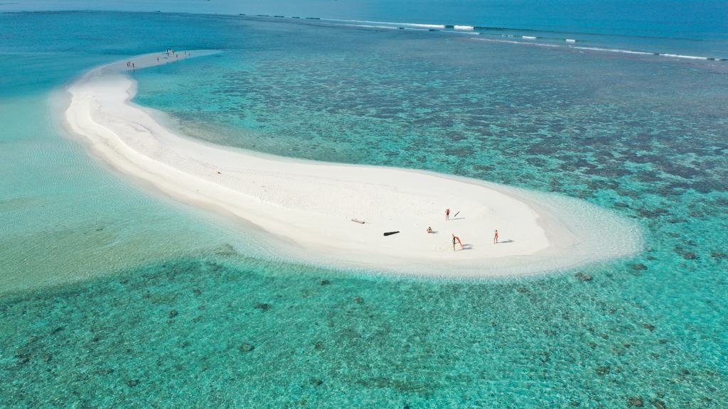 A sandbank from the air, ringed by pale shallows and dark reef, with a few figures standing on the sand.