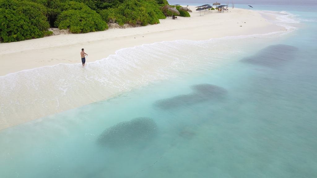Ukulhas beach from the air: one person standing in the shallows, parasols under the treeline, coral heads dark in the lagoon.
